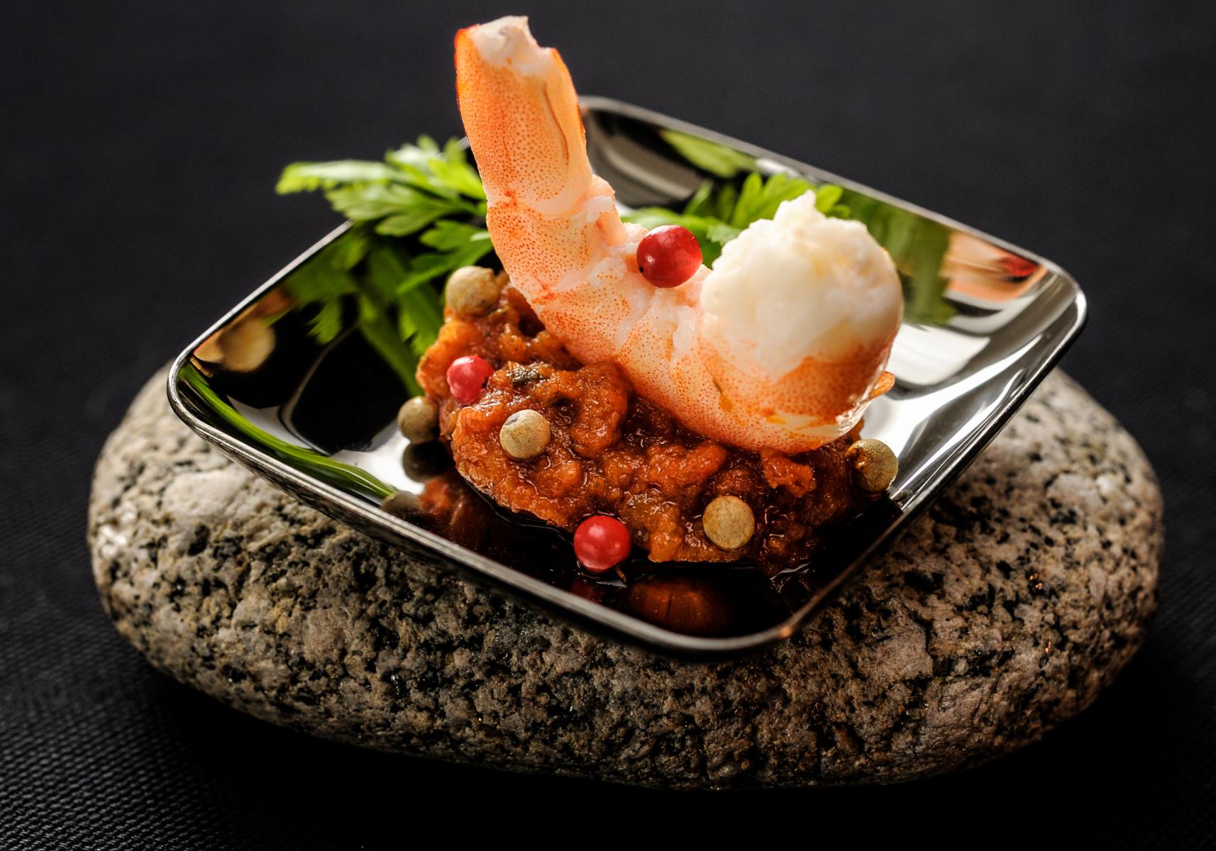 Gambas dressée sur une compotée d’épices et baies roses dans une coupelle noire posée sur un galet brut.