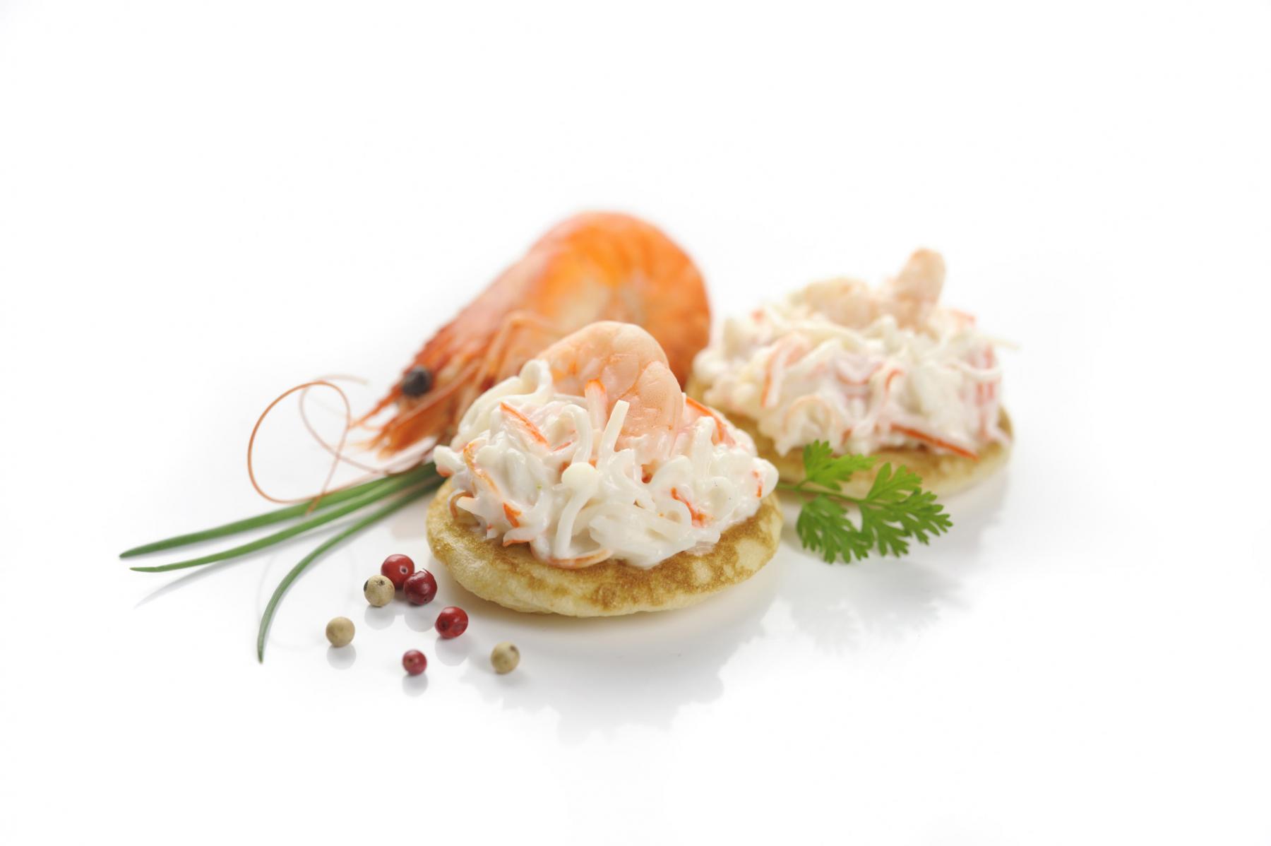 Deux mini blinis garnis de crème de surimi et crevettes avec baies roses sur fond blanc lumineux.