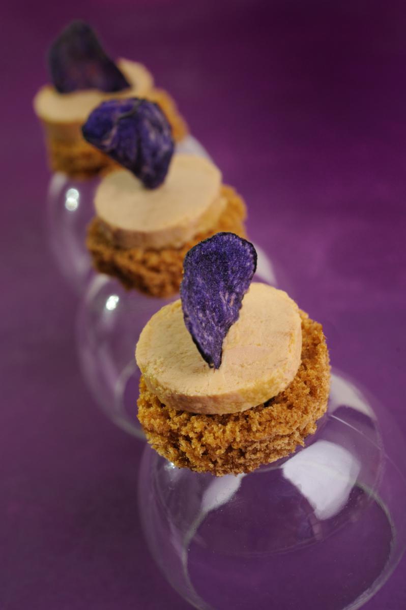 Toasts ronds de pain d’épices surmontés de foie gras et d’une chips de pomme de terre vitelotte sur fond violet.