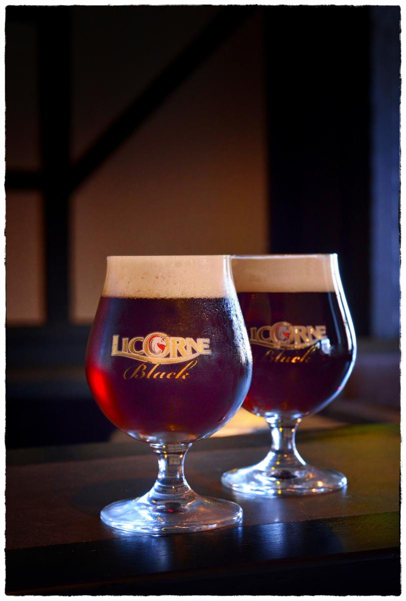 Deux verres ballon de bière brune Licorne Black avec une mousse onctueuse sous un éclairage clair-obscur en studio.