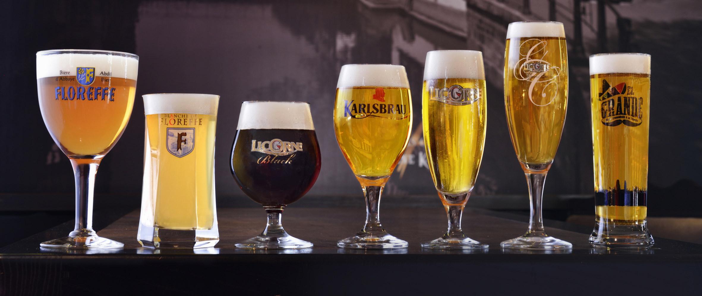 Sept verres de bières blondes, blanches et brunes avec mousse onctueuse, alignés sur une table sombre en studio.
