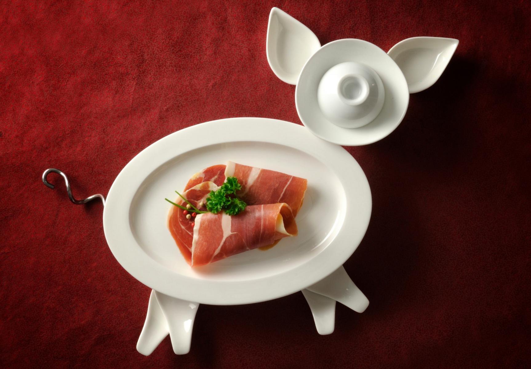 Assiettes en porcelaine blanche formant un cochon sur fond rouge avec tranches de jambon cru et persil en studio.