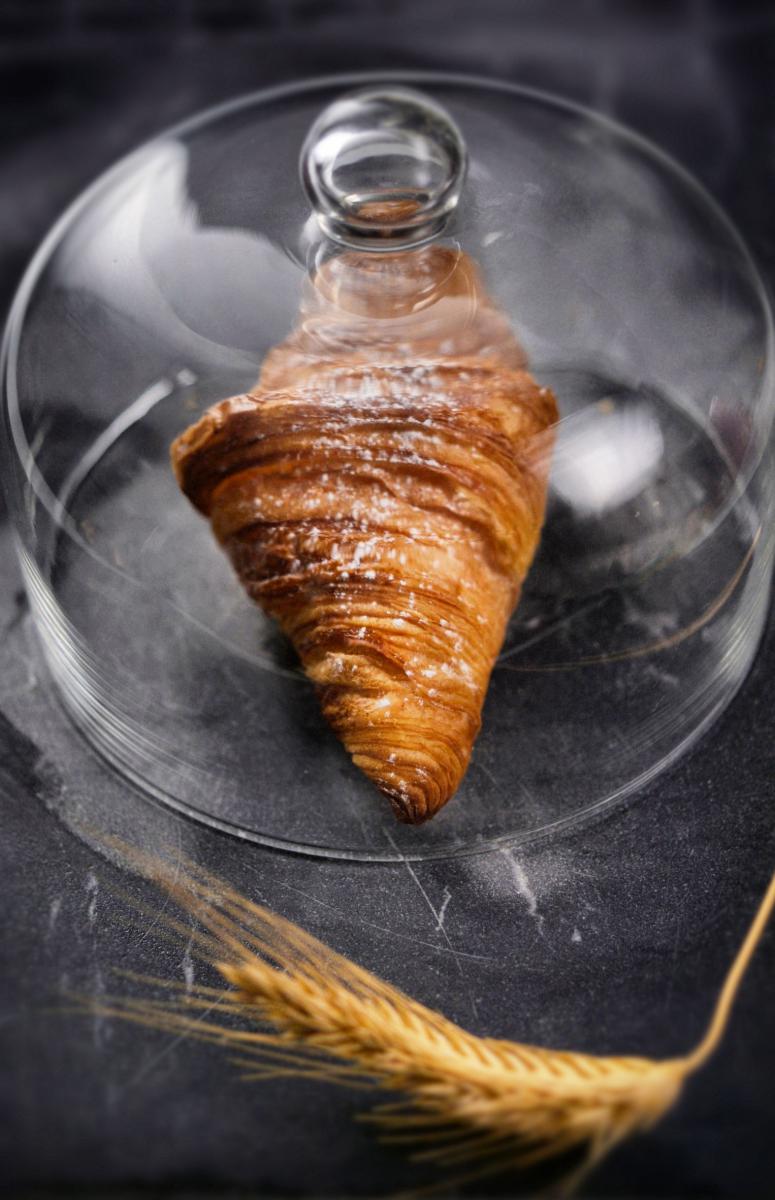 Croissant doré au feuilletage croustillant sous une cloche en verre sur marbre sombre avec un épi de blé.
