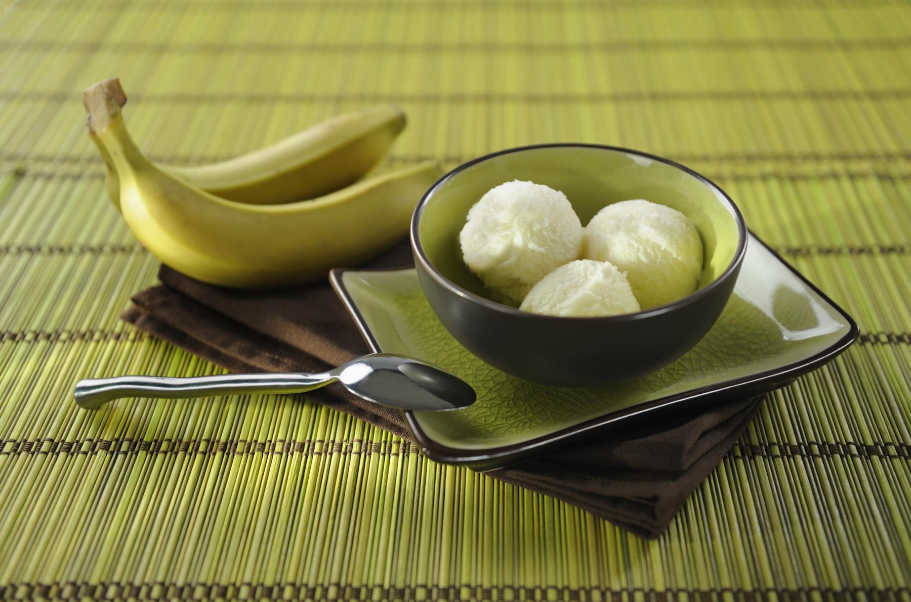 Trois boules de glace à la banane dans un bol sombre posé sur un set en bambou vert avec deux bananes entières.