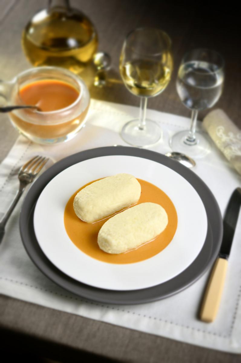 Quenelles sur sauce Nantua orange dans une assiette blanche, verres de vin blanc et saucière en arrière-plan lumineux.