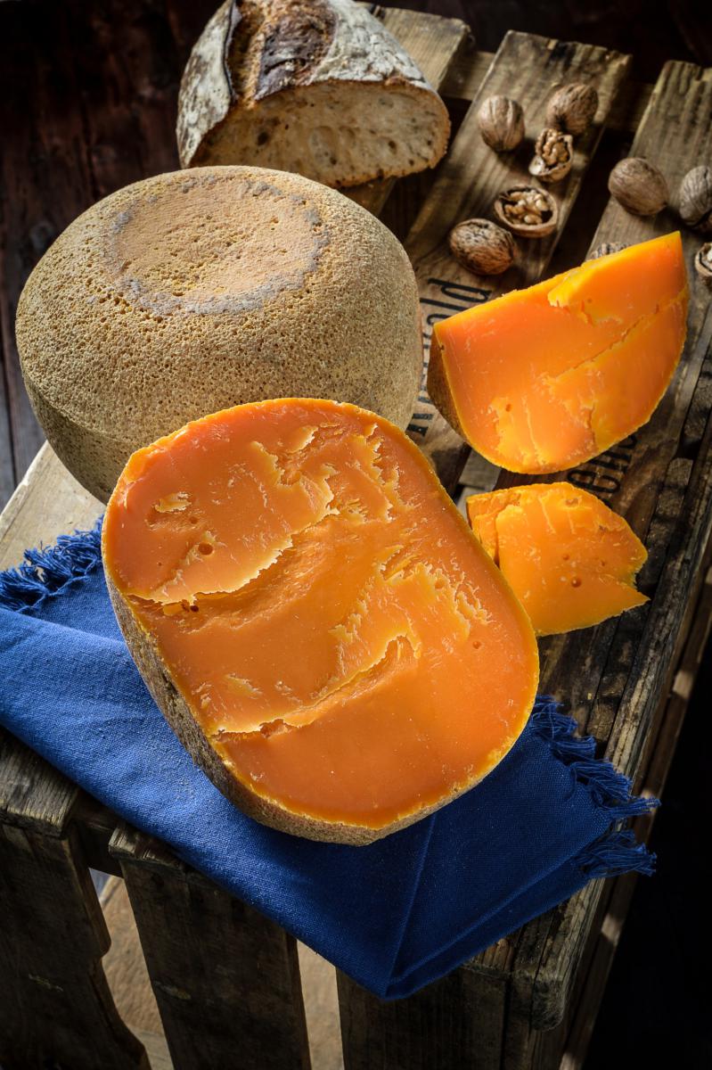 Meule de mimolette entamée révélant un cœur orange vif sur un cageot en bois avec du pain, des noix et un tissu bleu.