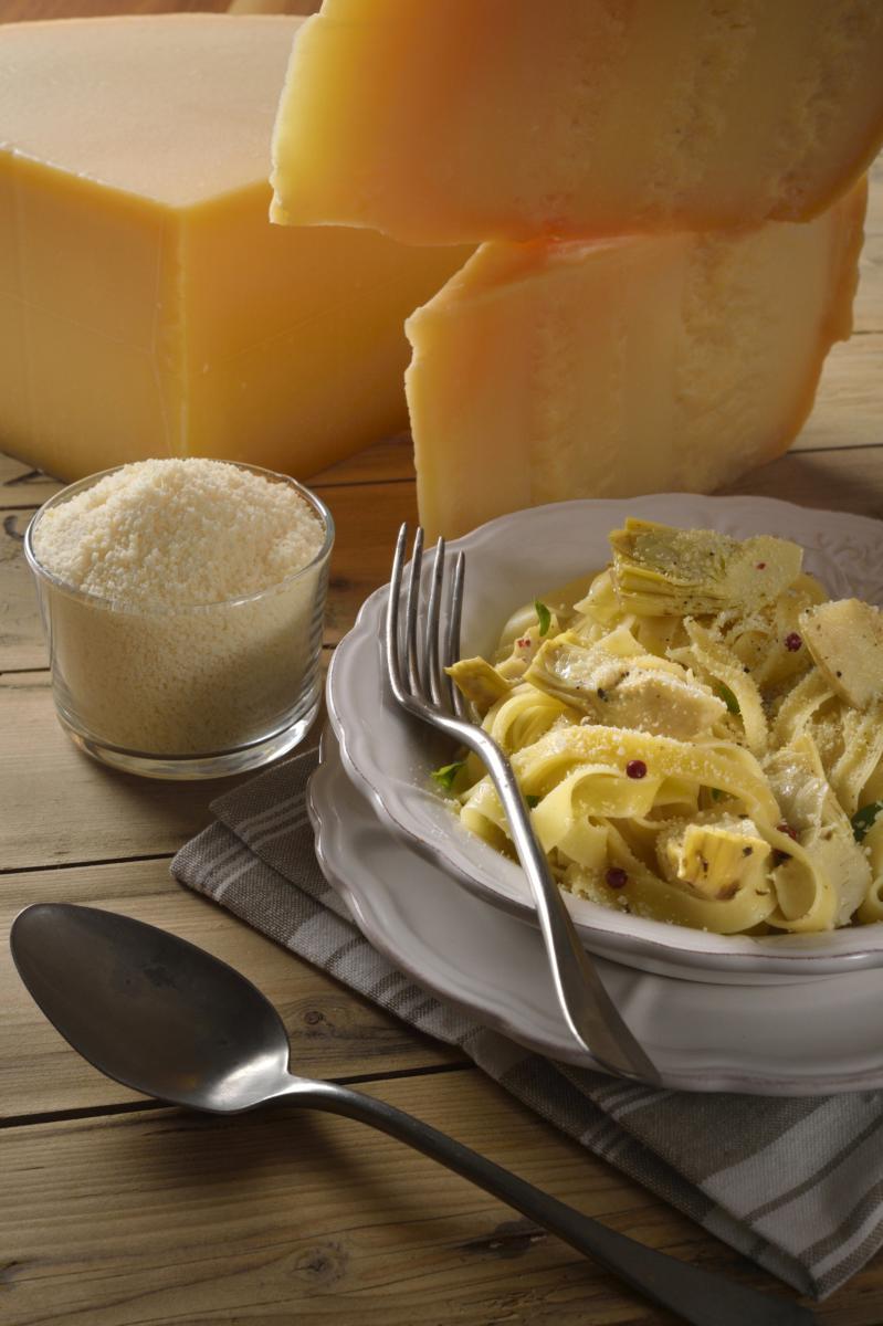 Assiette de pâtes aux artichauts et parmesan râpé sur une table en bois avec de larges blocs de fromage en arrière-plan.