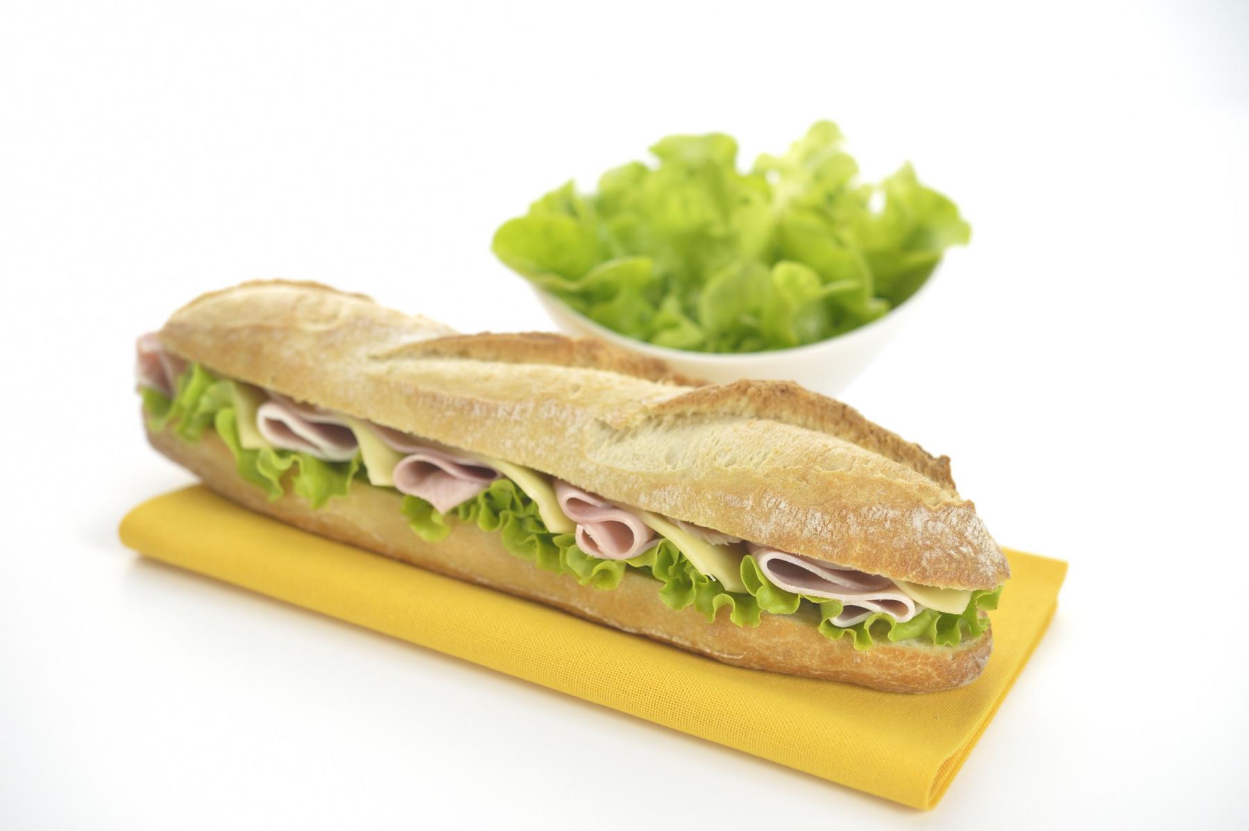 Sandwich baguette garni de jambon, emmental et salade sur serviette jaune, fond blanc lumineux, bol de salade verte.