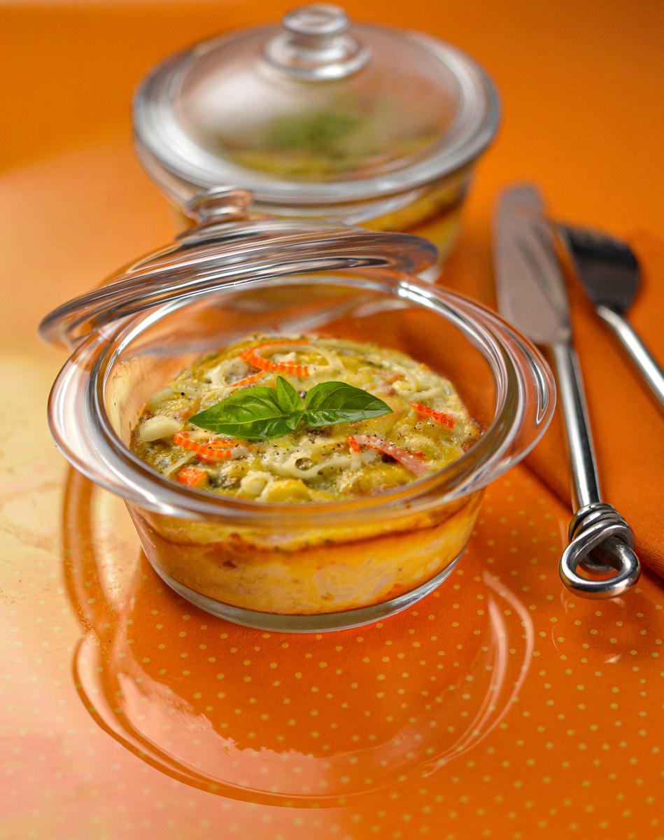 Cassolette de surimi gratinée dans un ramequin en verre avec feuille de basilic sur un fond orange et couverts en métal.