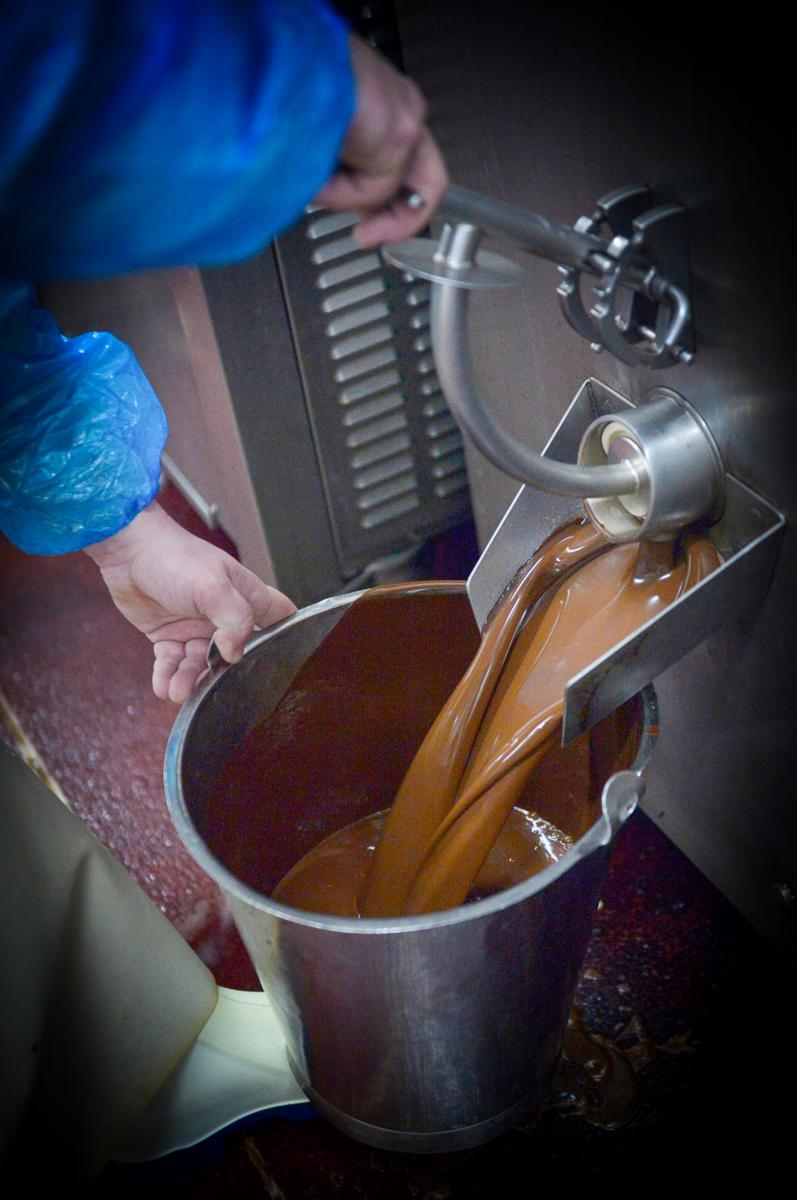 Coulée de chocolat liquide sortant d’une machine industrielle vers un seau en inox tenu par un opérateur en usine.