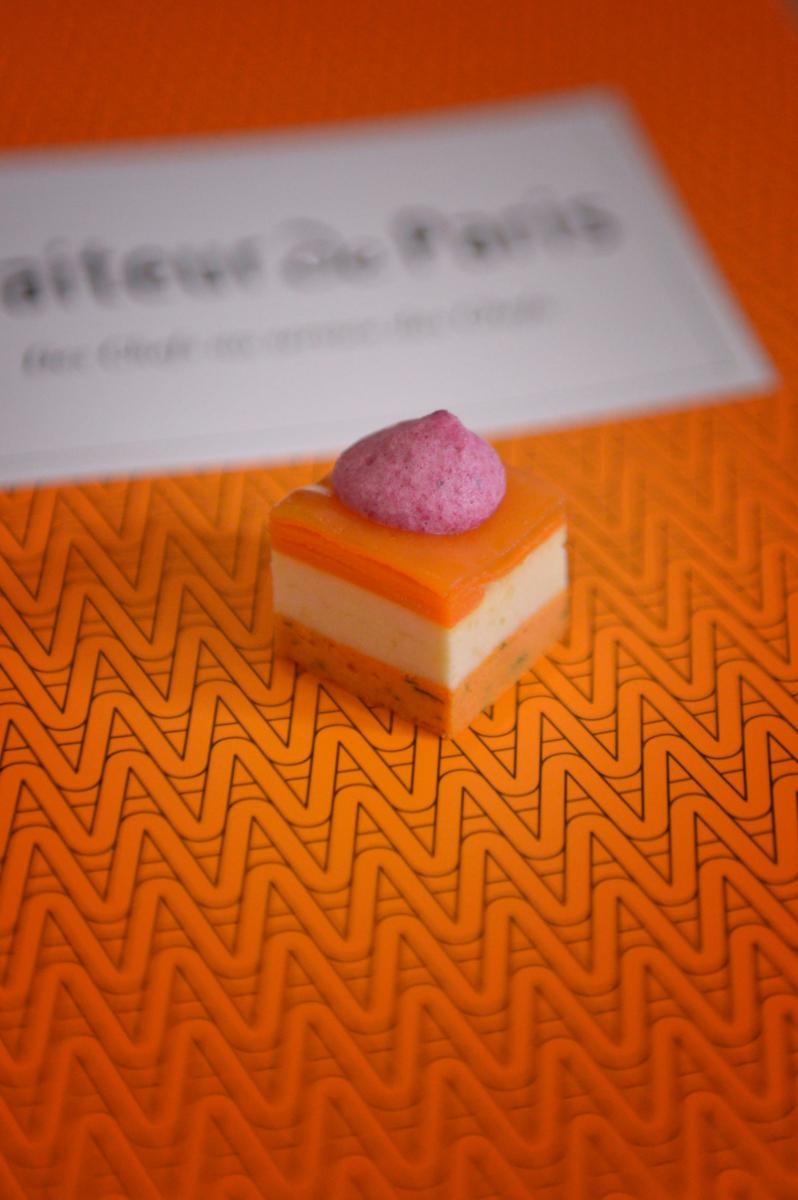 Mignardise cocktail carrée orange et blanche surmontée d’une mousse violette sur fond graphique orange vif