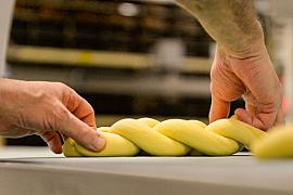 Mains d’un boulanger tressant une pâte à brioche jaune sur un plan de travail en inox sous un éclairage studio doux.