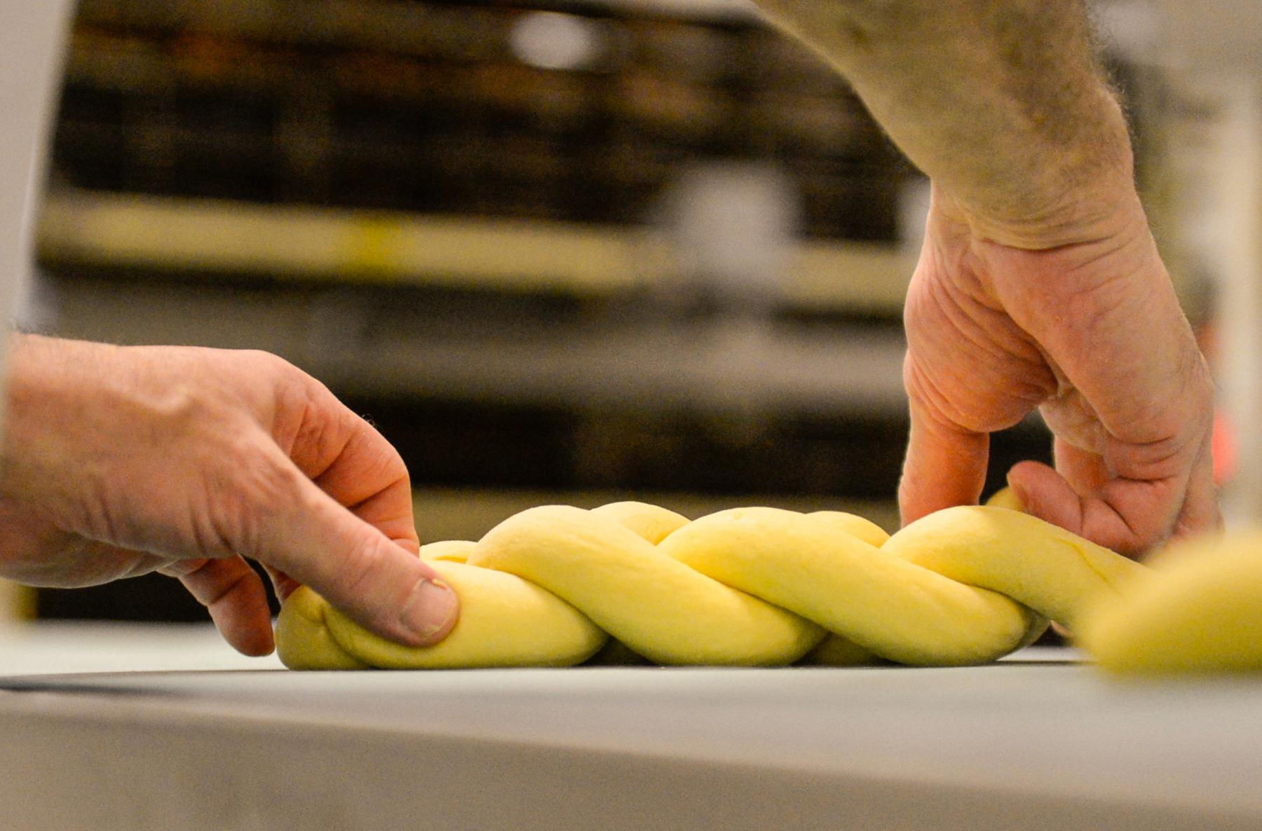 Mains d’un boulanger tressant une pâte à brioche jaune sur un plan de travail en inox sous un éclairage studio doux.