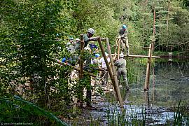 Groupe en tenue de travail construisant un pont laotien, de bois et cordes au-dessus d’un étang forestier.