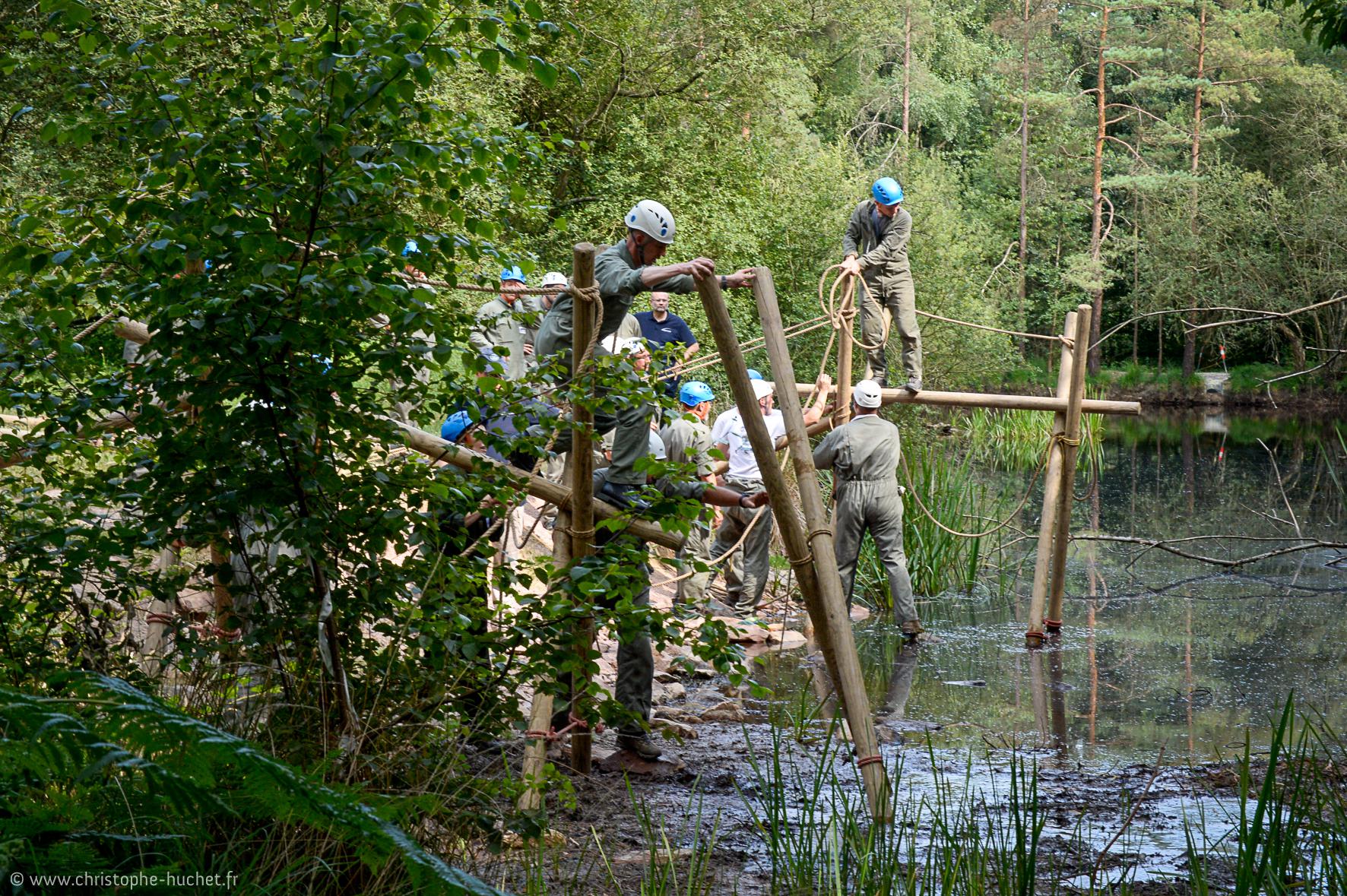 Groupe en tenue de travail construisant un pont laotien, de bois et cordes au-dessus d’un étang forestier.