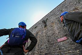 Deux hommes casqués observent un grimpeur sur un haut mur de pierre sous un ciel bleu lors d’un exercice de groupe.