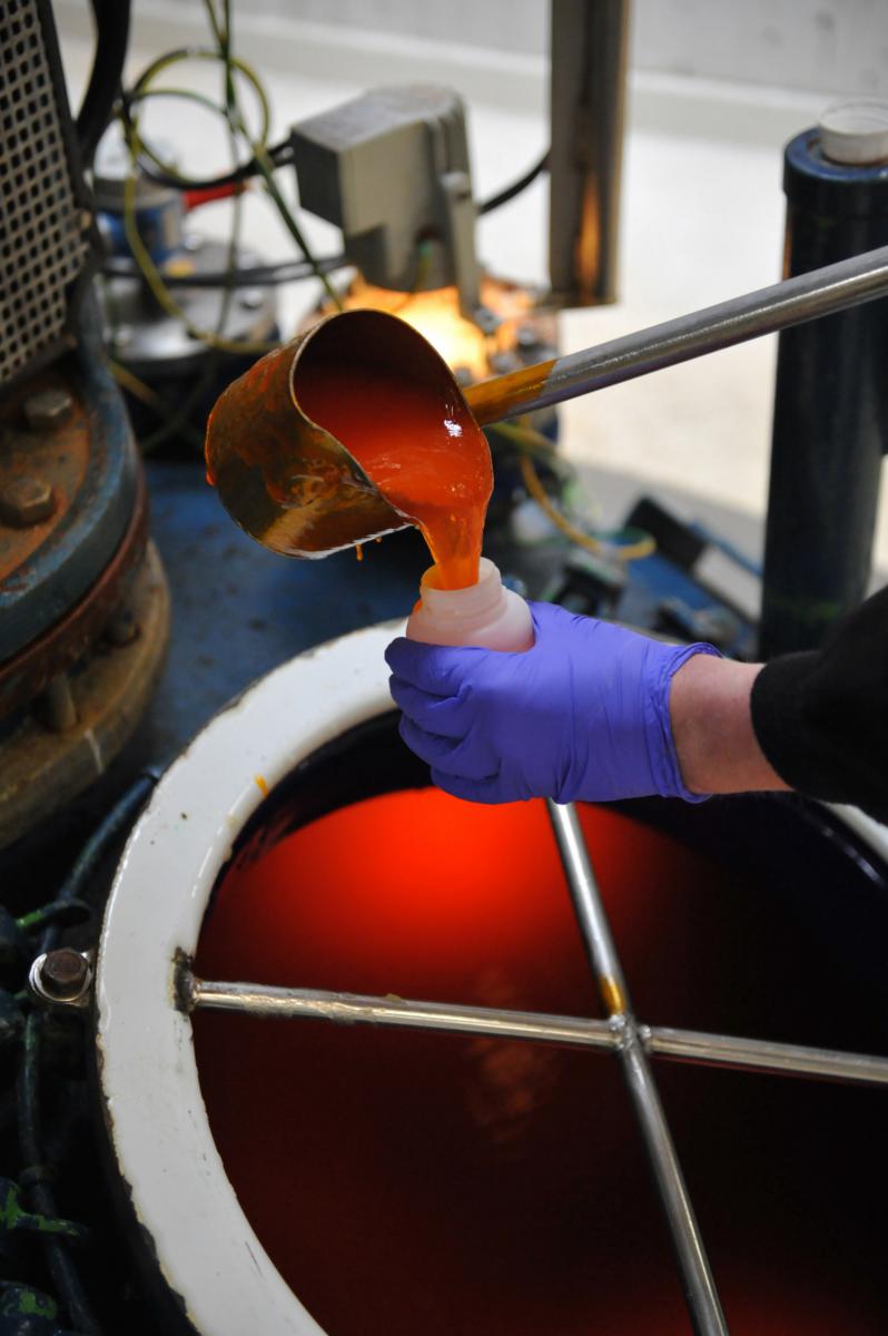 Main gantée versant un liquide orange vif dans un flacon au-dessus d’une cuve industrielle éclairée