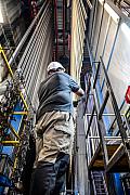 Ouvrier casqué au pied de rails verticaux de peinture industrielle dans une usine bretonne sous un éclairage zénithal.