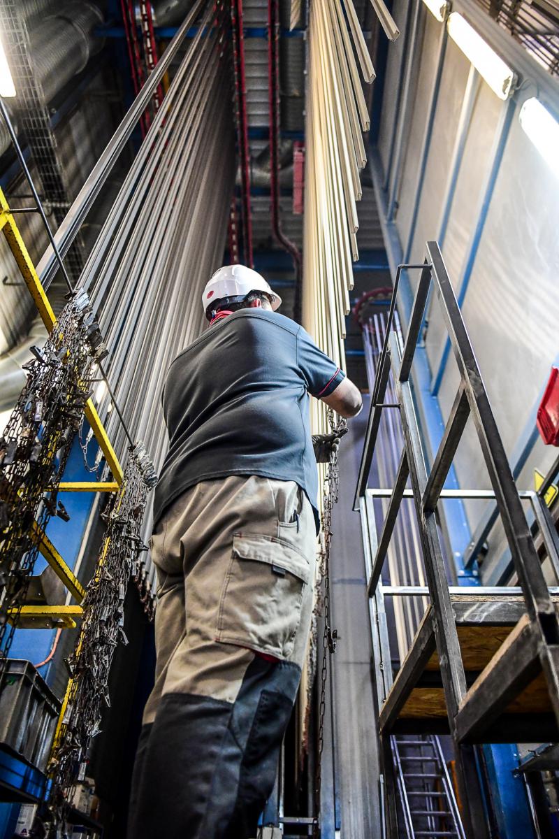 Ouvrier casqué au pied de rails verticaux de peinture industrielle dans une usine bretonne sous un éclairage zénithal.