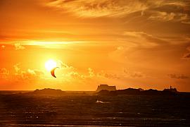 Silhouette d’aile de kitesurf devant un grand soleil couchant orange, mer agitée et forts de Saint-Malo à l’horizon.