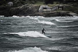 Silhouette d’un surfeur en paddle sur une vague blanche devant une côte rocheuse et un bunker gris en Bretagne.