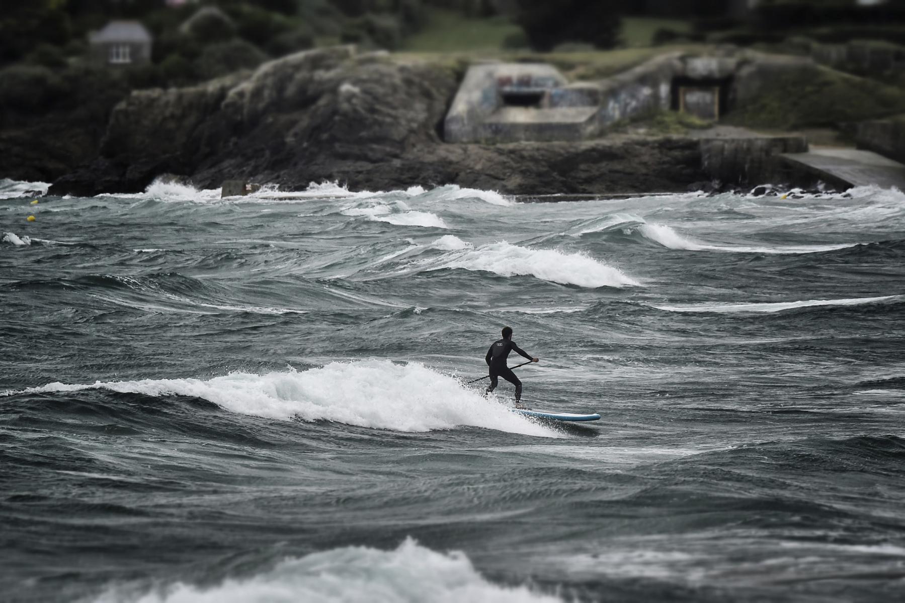 Silhouette d’un surfeur en paddle sur une vague blanche devant une côte rocheuse et un bunker gris en Bretagne.