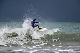 Surfeur en combinaison noire sur une vague déferlante avec éclaboussures d’écume sous un ciel sombre et menaçant.