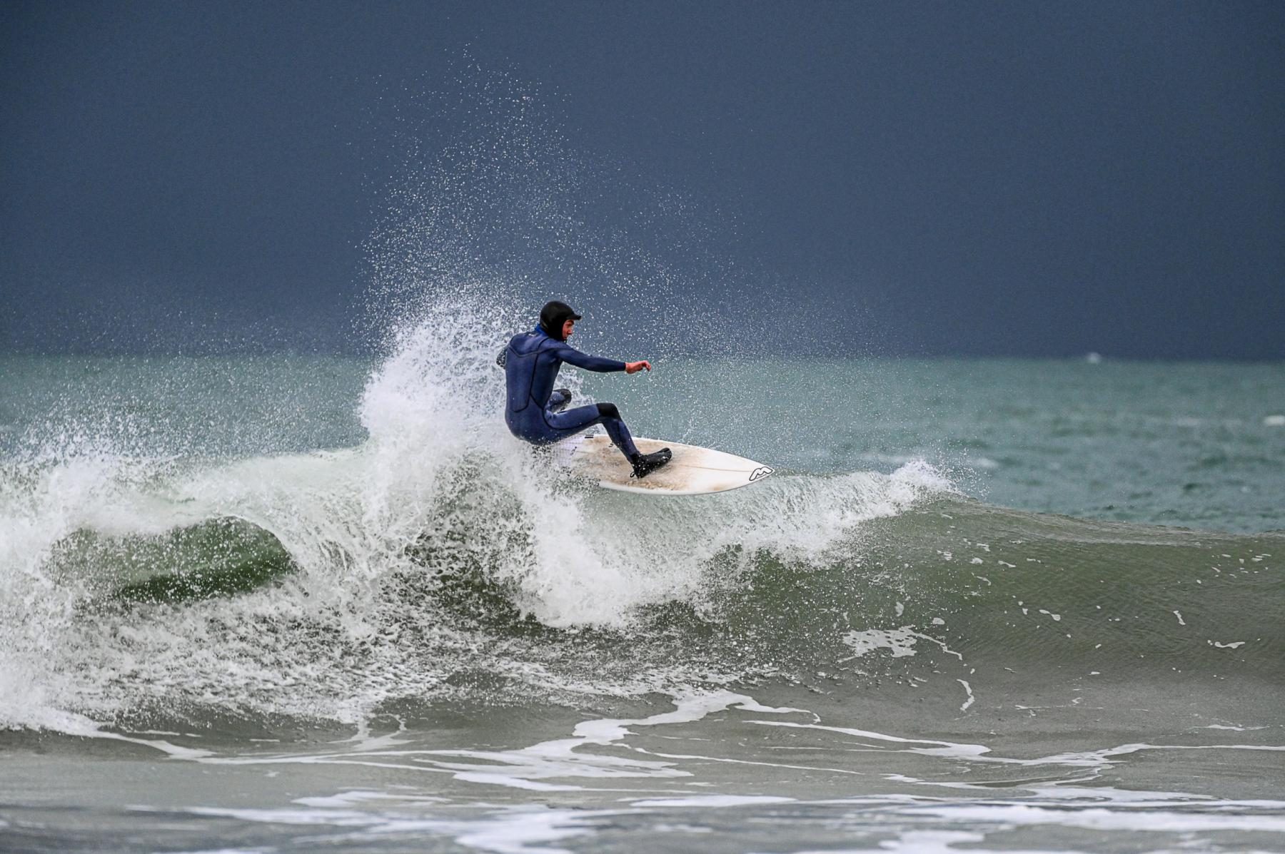 Surfeur en combinaison noire sur une vague déferlante avec éclaboussures d’écume sous un ciel sombre et menaçant.