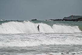 Surfeur en noir sur une vague blanche à Saint-Lunaire, mer grise et côte rocheuse en fond.