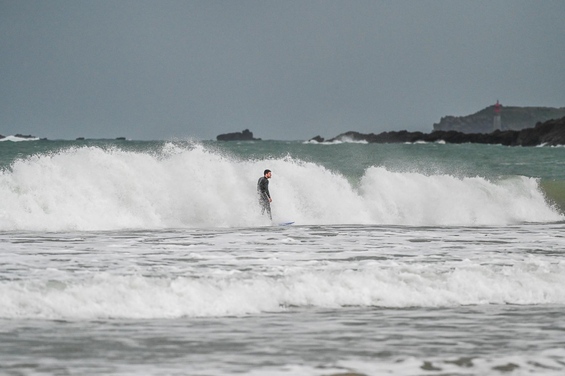 Surfeur en noir sur une vague blanche à Saint-Lunaire, mer grise et côte rocheuse en fond.