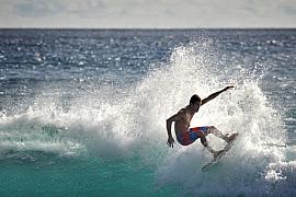 Surfeur en équilibre sur une crête de vague déferlante avec projections d’écume blanche sous un soleil intense.