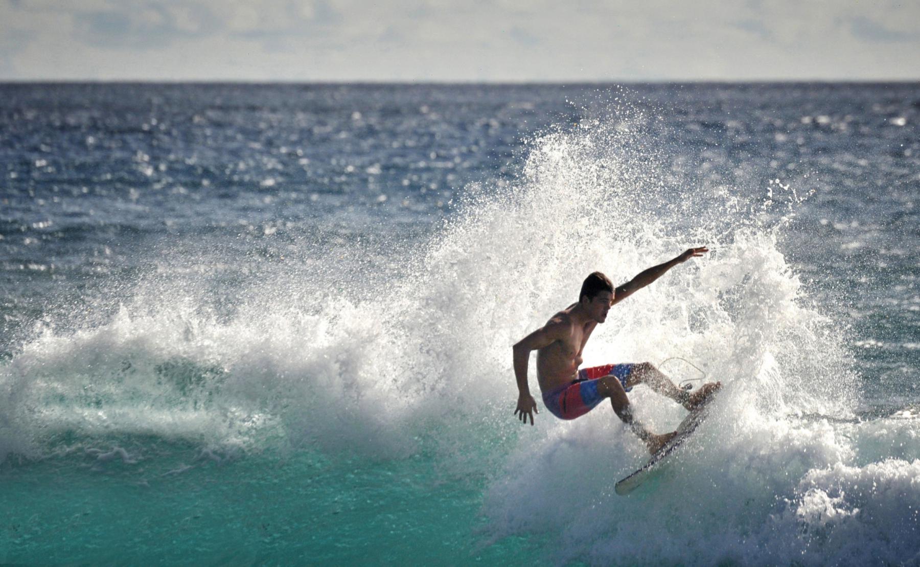 Surfeur en équilibre sur une crête de vague déferlante avec projections d’écume blanche sous un soleil intense.