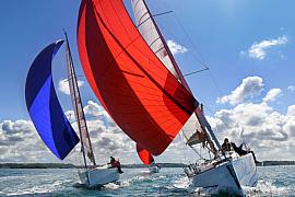 Deux voiliers avec spinnakers rouge et bleu naviguant côte à côte sur une mer calme sous un ciel ensoleillé