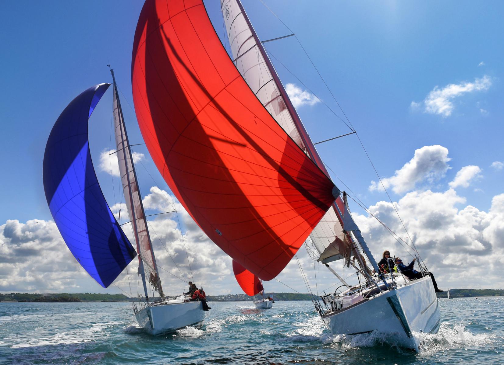 Deux voiliers avec spinnakers rouge et bleu naviguant côte à côte sur une mer calme sous un ciel ensoleillé