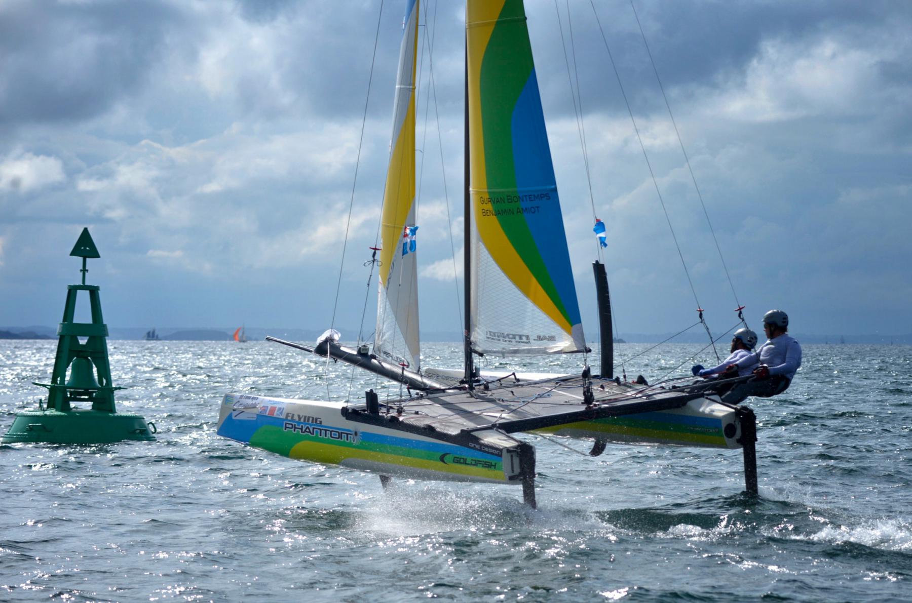 Catamaran volant sur ses foils, voiles colorées, deux skippeurs en rappel sur une mer agitée avec une balise verte au loin