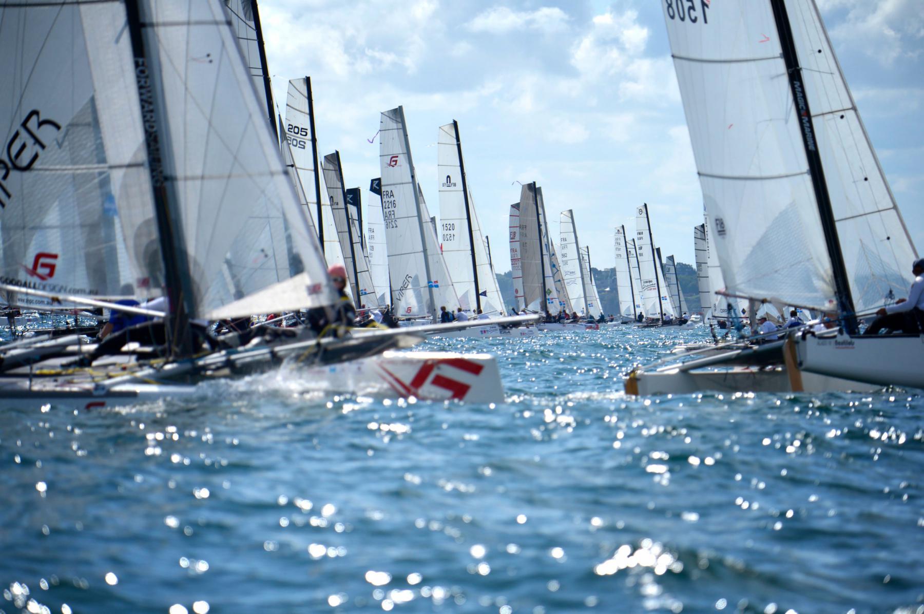 Multiples voiles de catamarans en mer. Lumière vive, reflets sur l’eau et départ de course nautique.