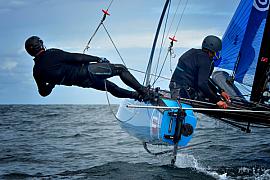 Deux navigateurs sur un catamaran bleu à foils en mer, un homme en noir au trapèze au-dessus de l’eau.