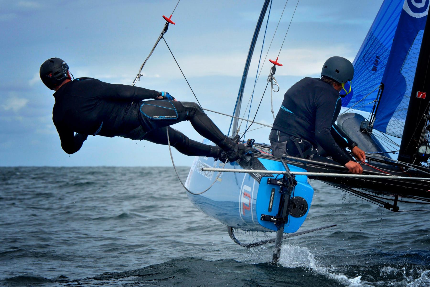 Deux navigateurs sur un catamaran bleu à foils en mer, un homme en noir au trapèze au-dessus de l’eau.