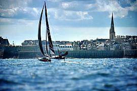 Voilier catamaran à foils en navigation rapide devant les remparts et le clocher de la ville de Saint-Malo.