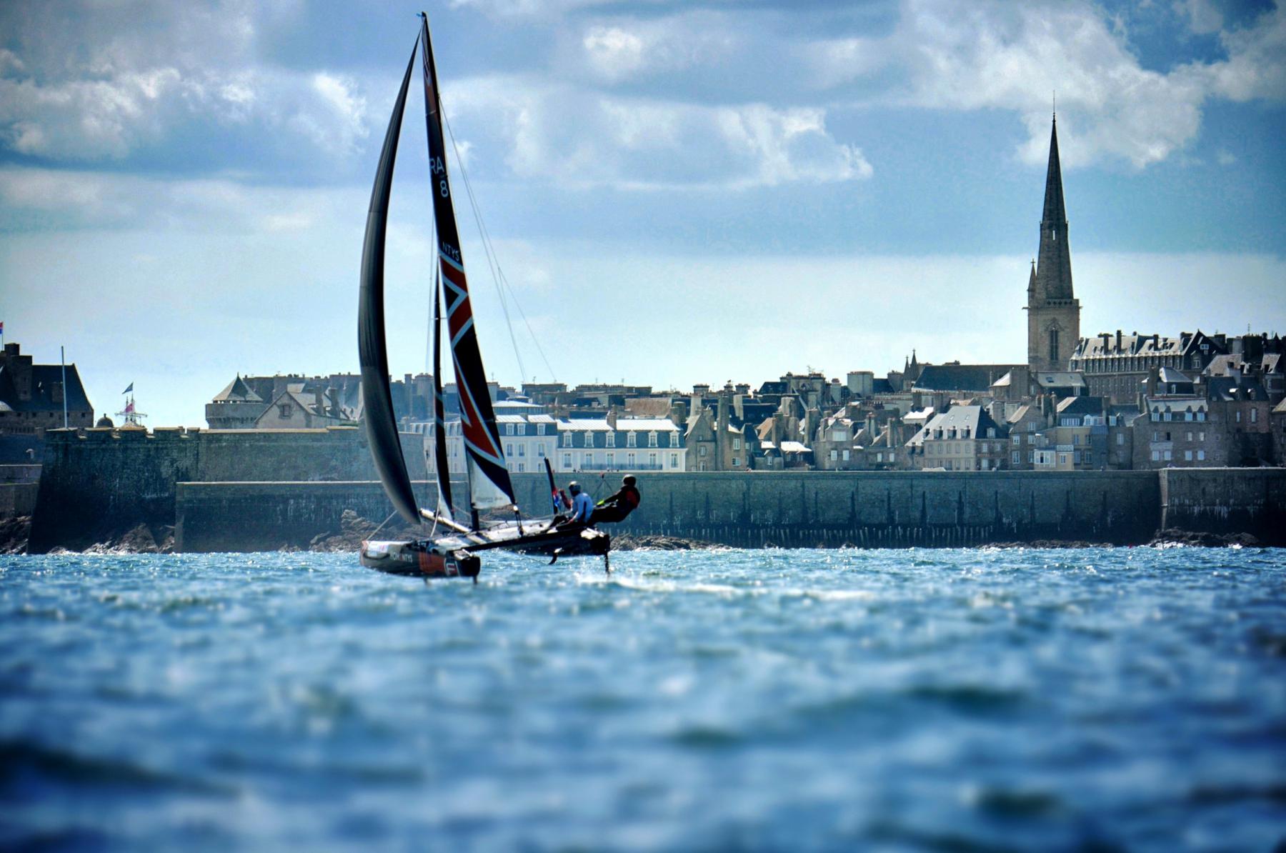 Voilier catamaran à foils en navigation rapide devant les remparts et le clocher de la ville de Saint-Malo.