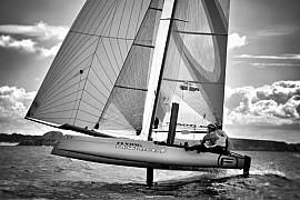 Catamaran Flying Phantom en noir et blanc sur ses foils au-dessus de l’eau avec deux skippers en pleine vitesse.