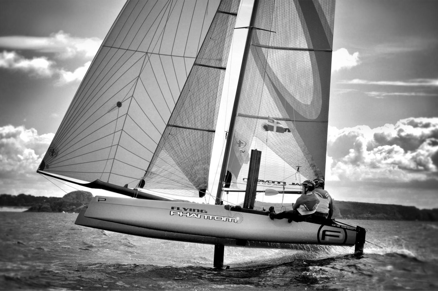 Catamaran Flying Phantom en noir et blanc sur ses foils au-dessus de l’eau avec deux skippers en pleine vitesse.