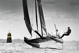 Catamaran à foils s’élevant au-dessus de l’eau, équipage en action sous un ciel nuageux près d’une balise maritime.