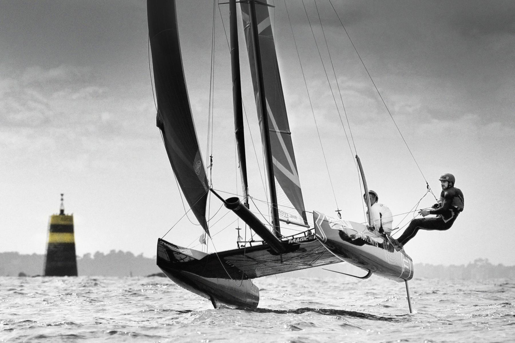Catamaran à foils s’élevant au-dessus de l’eau, équipage en action sous un ciel nuageux près d’une balise maritime.