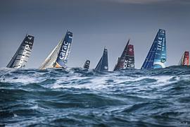 Voiliers Class 40 aux voiles colorées franchissant une vague d’écume blanche dans la houle sous un ciel gris menaçant.