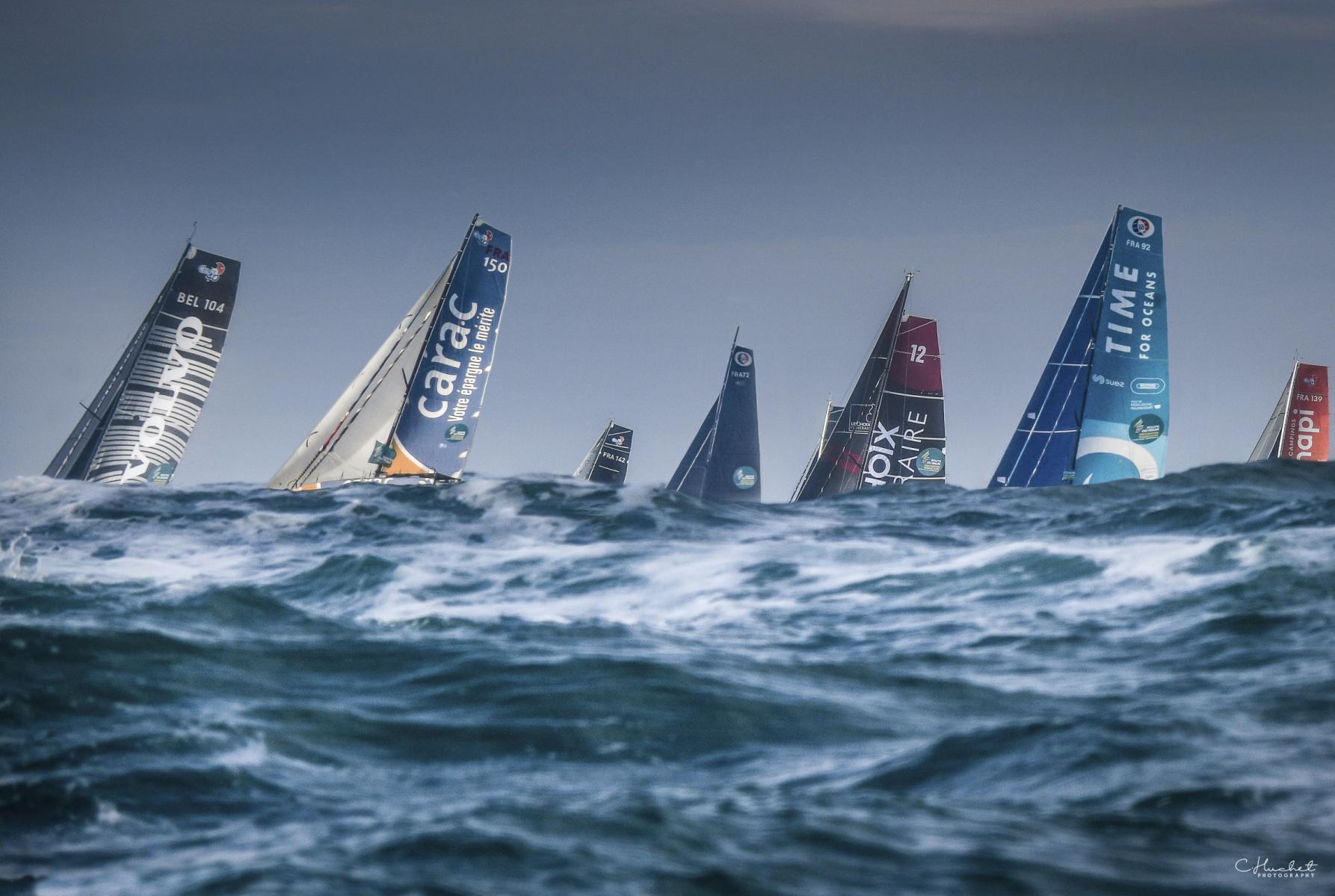 Voiliers Class 40 aux voiles colorées franchissant une vague d’écume blanche dans la houle sous un ciel gris menaçant.
