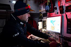 Skipper de profil avec lampe frontale scrutant un écran de navigation dans la cabine sombre d’un voilier.