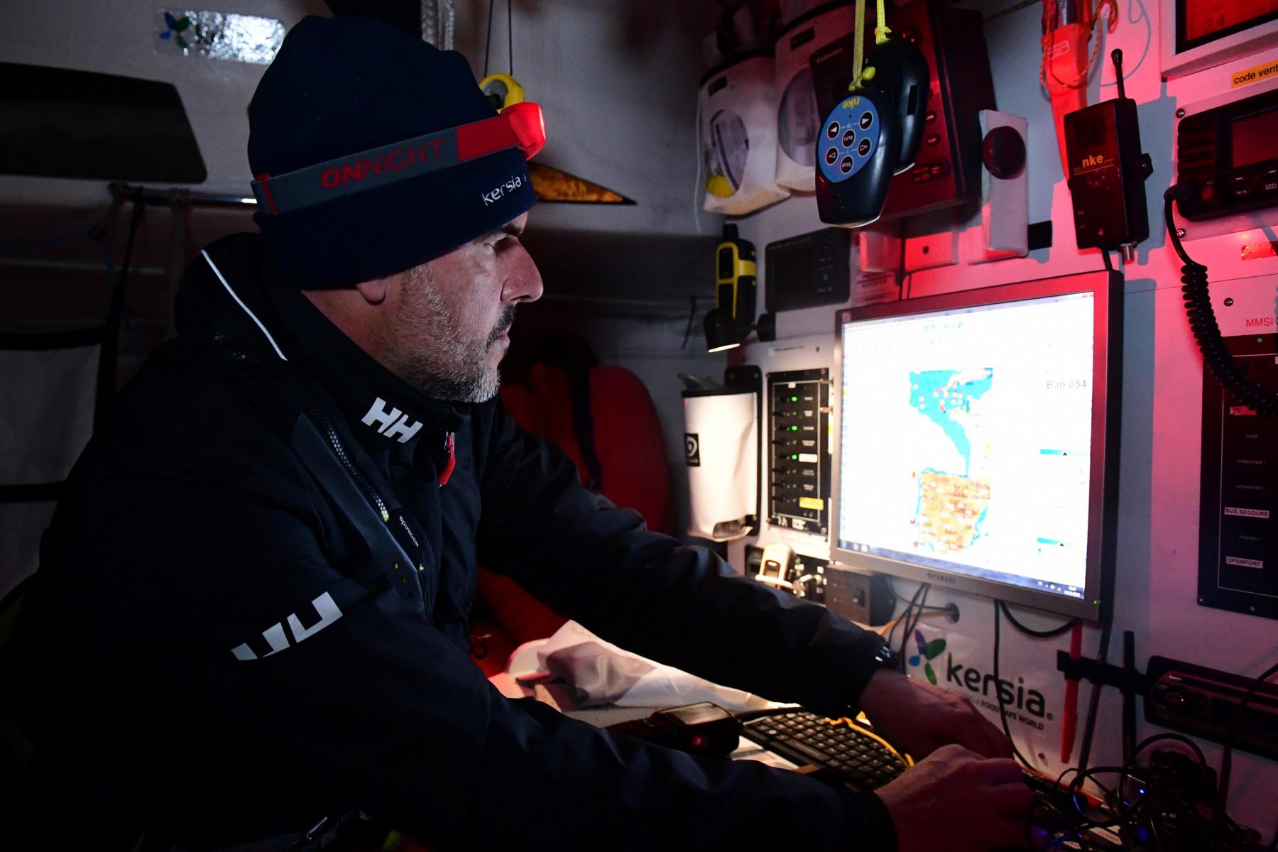 Skipper de profil avec lampe frontale scrutant un écran de navigation dans la cabine sombre d’un voilier.