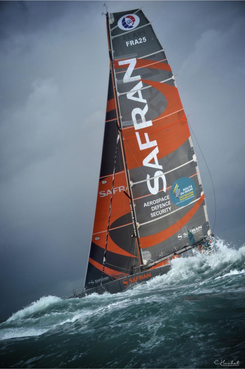 Voilier IMOCA Safran sur mer agitée avec vagues blanches et ciel sombre lors de la Route du Rhum à Saint-Malo.