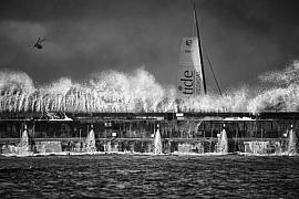 Voilier IMOCA derrière une jetée sous les embruns au Havre, hélicoptère en vol, photographie noir et blanc contrastée.